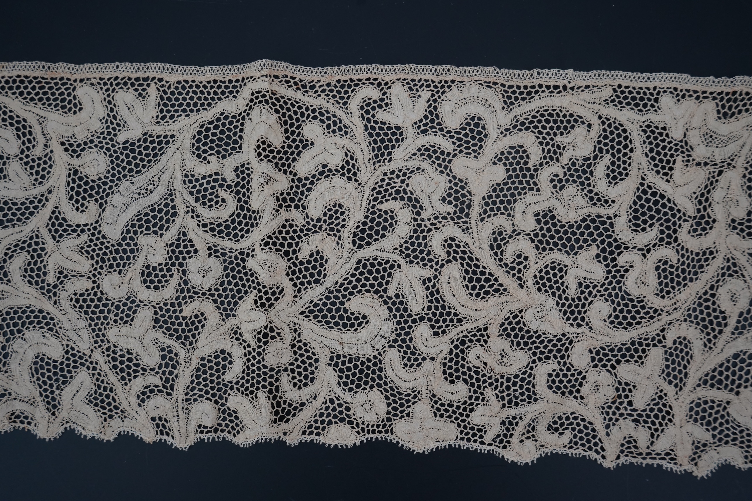 A length of 1740-1760 Italian, Milanese bobbin lace flounce, made from flat braiding or tapes, placed into scrolling patterns, attached to brides, made into a honeycomb mesh. 252cm long x 16cm deep. Condition - one small